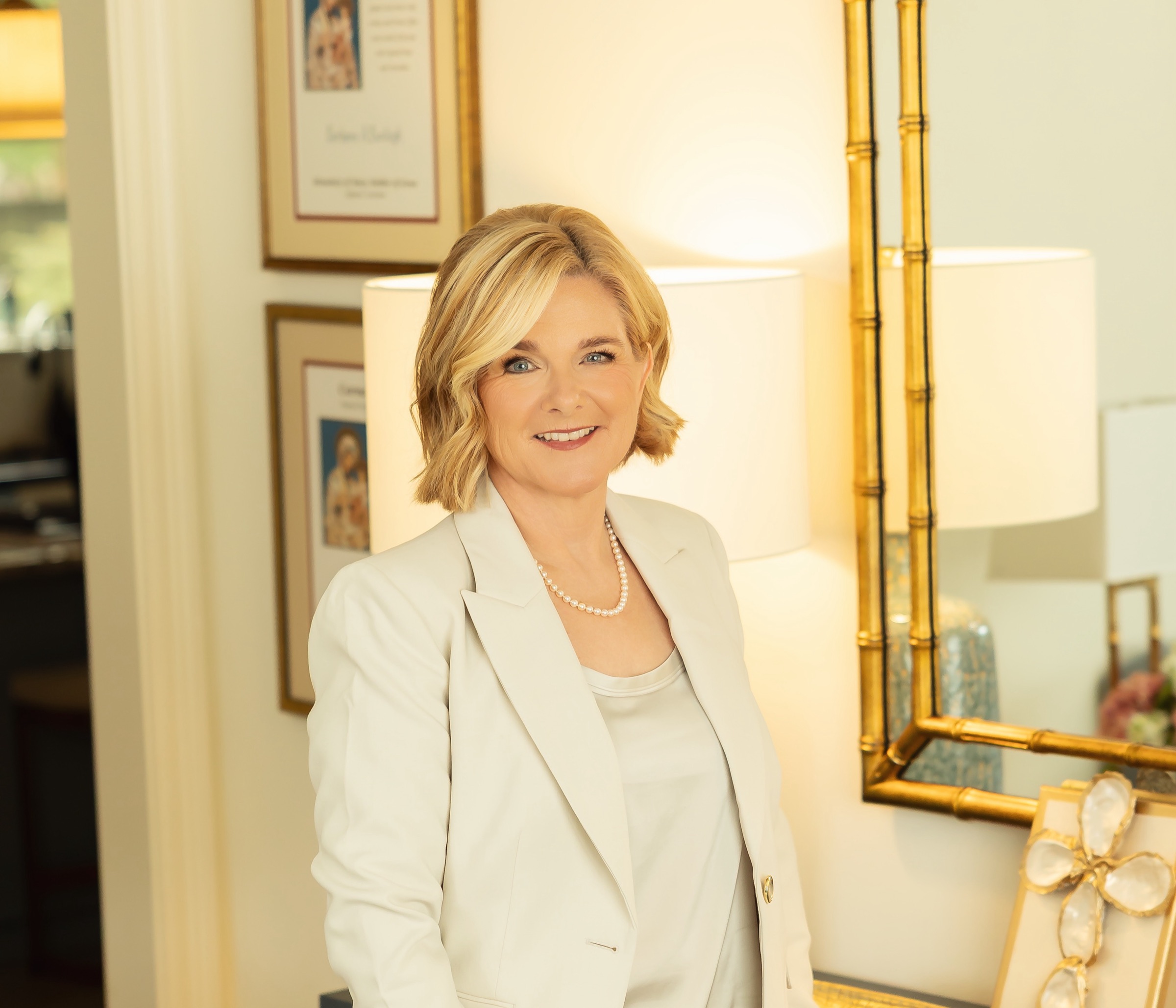 Lisa Romero Dugal beside a gilt mirror, smiling, in a cream suit