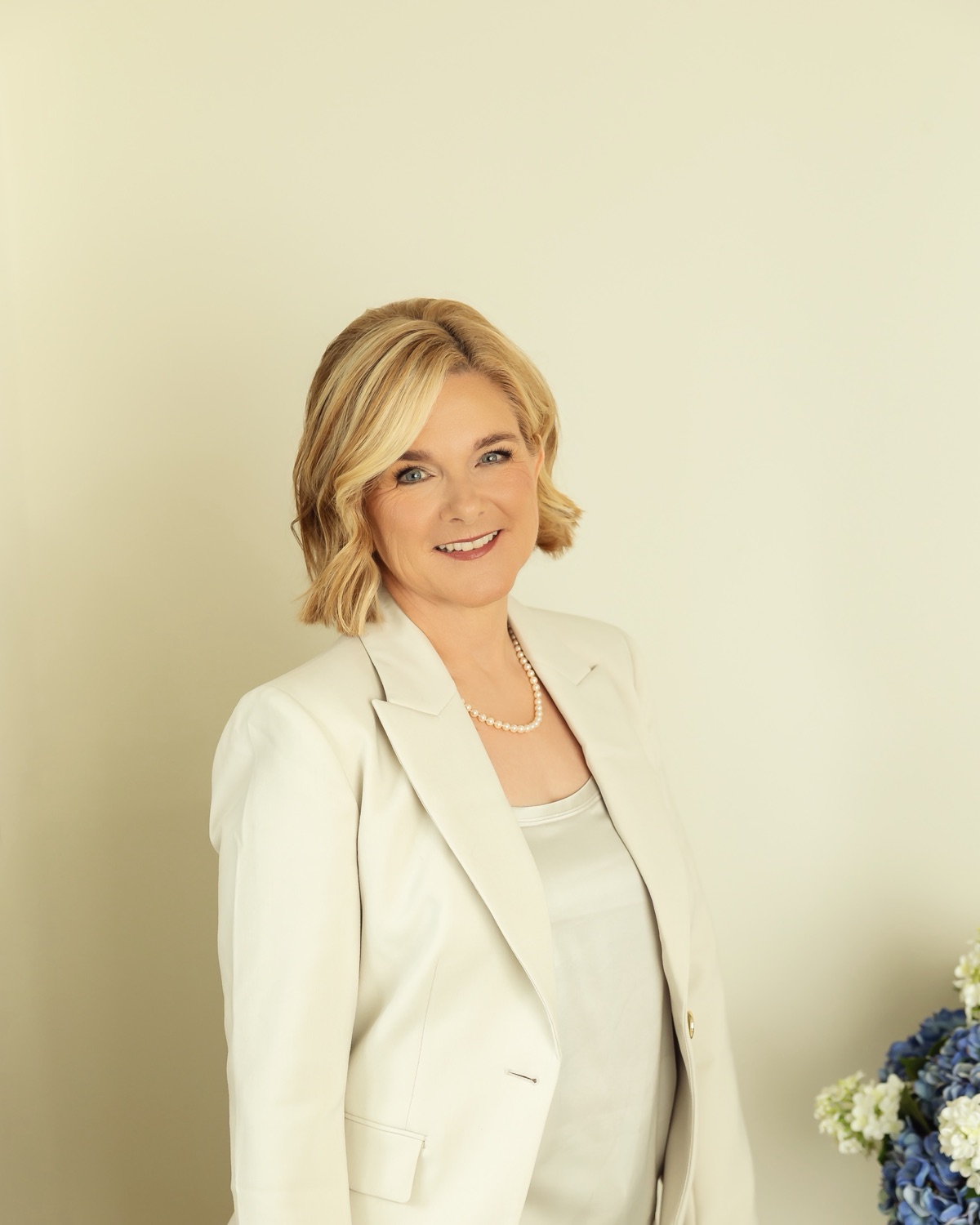 Lisa Romero Dugal in a cream suit against a cream wall, blue hydrangeas beside her